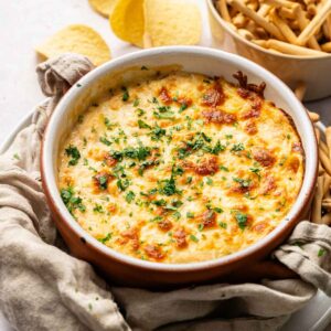 A round baking dish filled with cheesy Hot Crab Dip, garnished with chopped herbs, sits next to a bowl of breadsticks and several potato chips.
