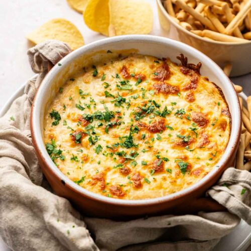 A round baking dish filled with cheesy Hot Crab Dip, garnished with chopped herbs, sits next to a bowl of breadsticks and several potato chips.