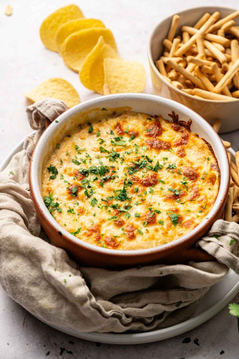 A round baking dish filled with cheesy Hot Crab Dip, garnished with chopped herbs, sits next to a bowl of breadsticks and several potato chips.