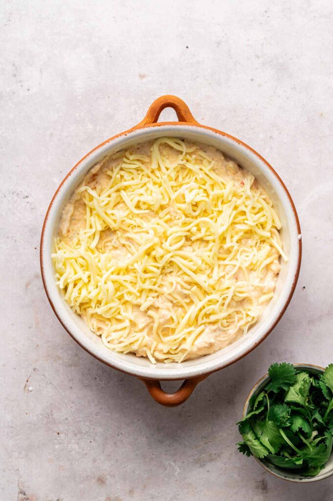 A round baking dish filled with unbaked Hot Crab Dip, topped with shredded cheese, sits on a light surface next to a small bowl of fresh cilantro.
