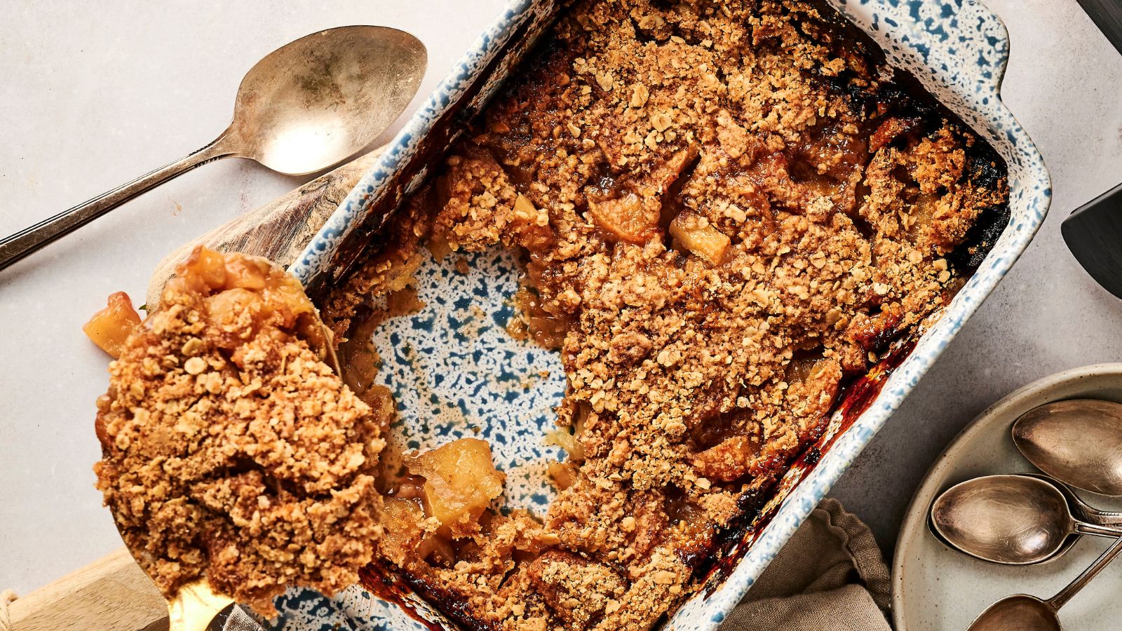 A baked apple crumble in a speckled baking dish with a serving spoon, some crumble scooped out, and extra spoons nearby.