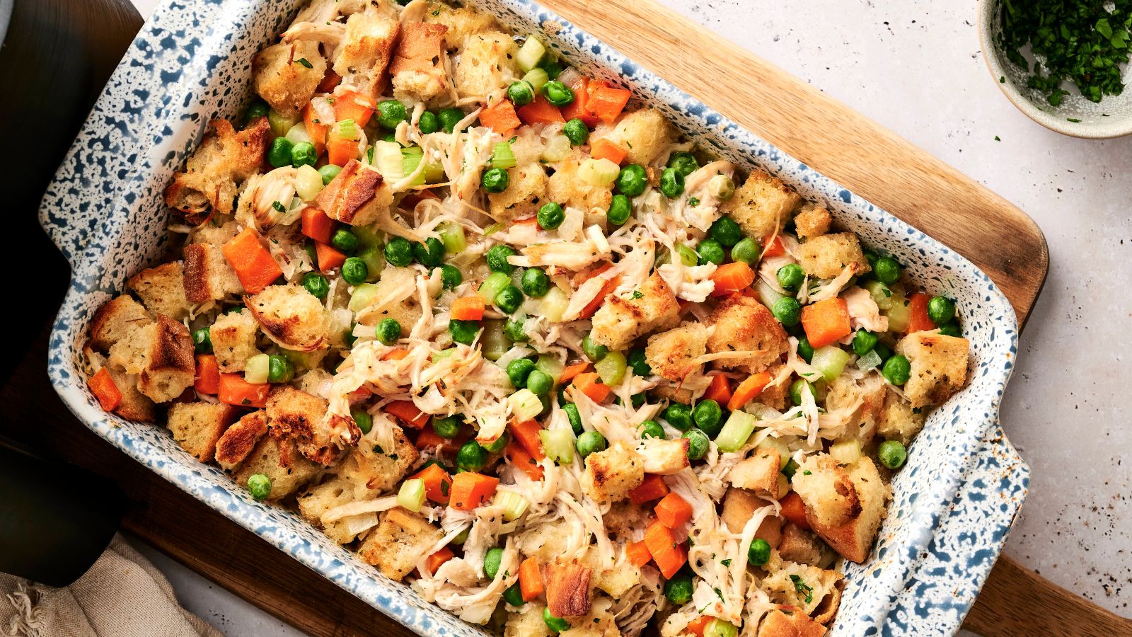 A casserole dish filled with baked bread cubes, shredded chicken, peas, carrots, and celery, arranged on a wooden board.