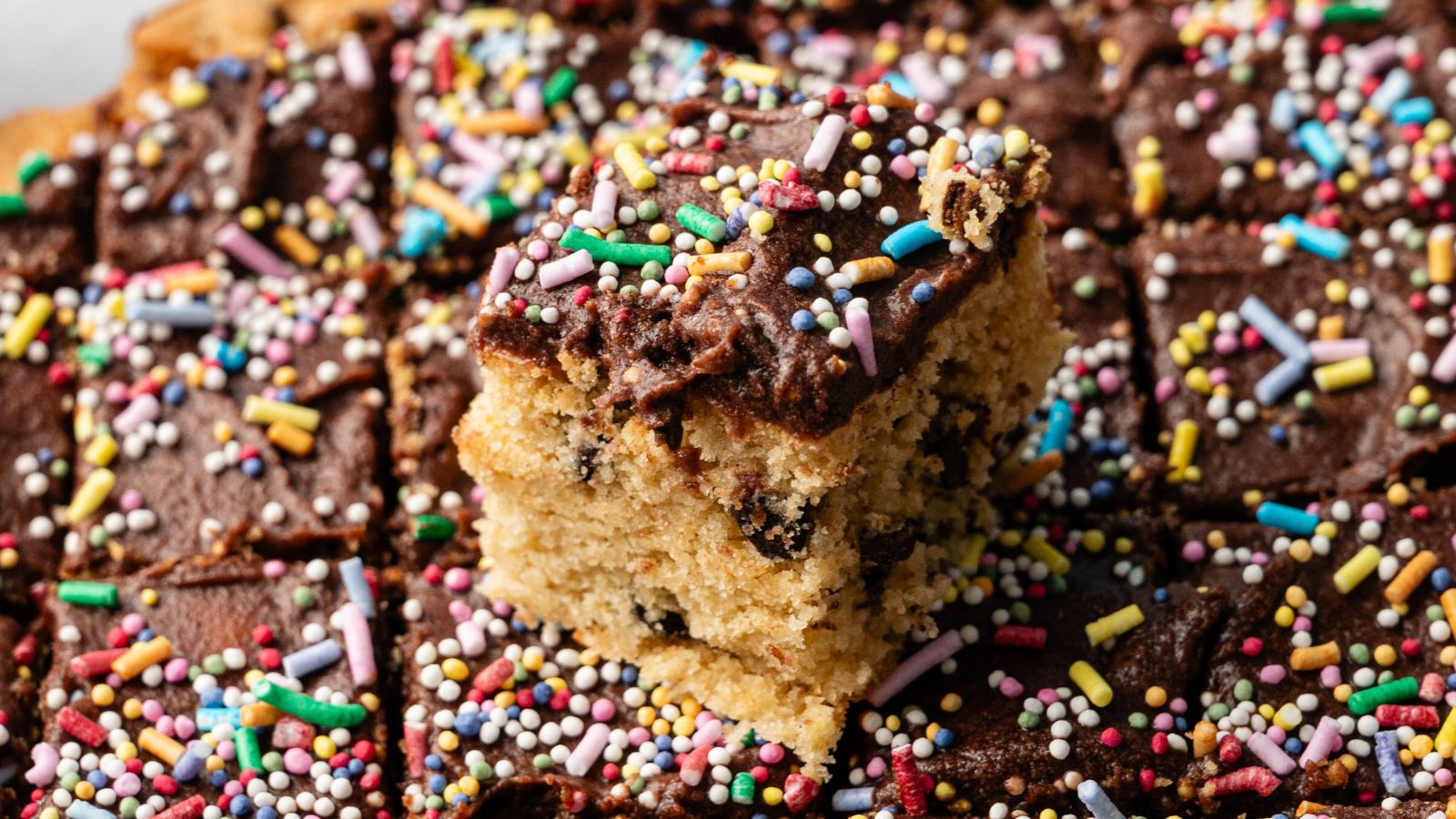 A tray of chocolate-frosted cake squares with colorful sprinkles, featuring one piece slightly raised above the rest.