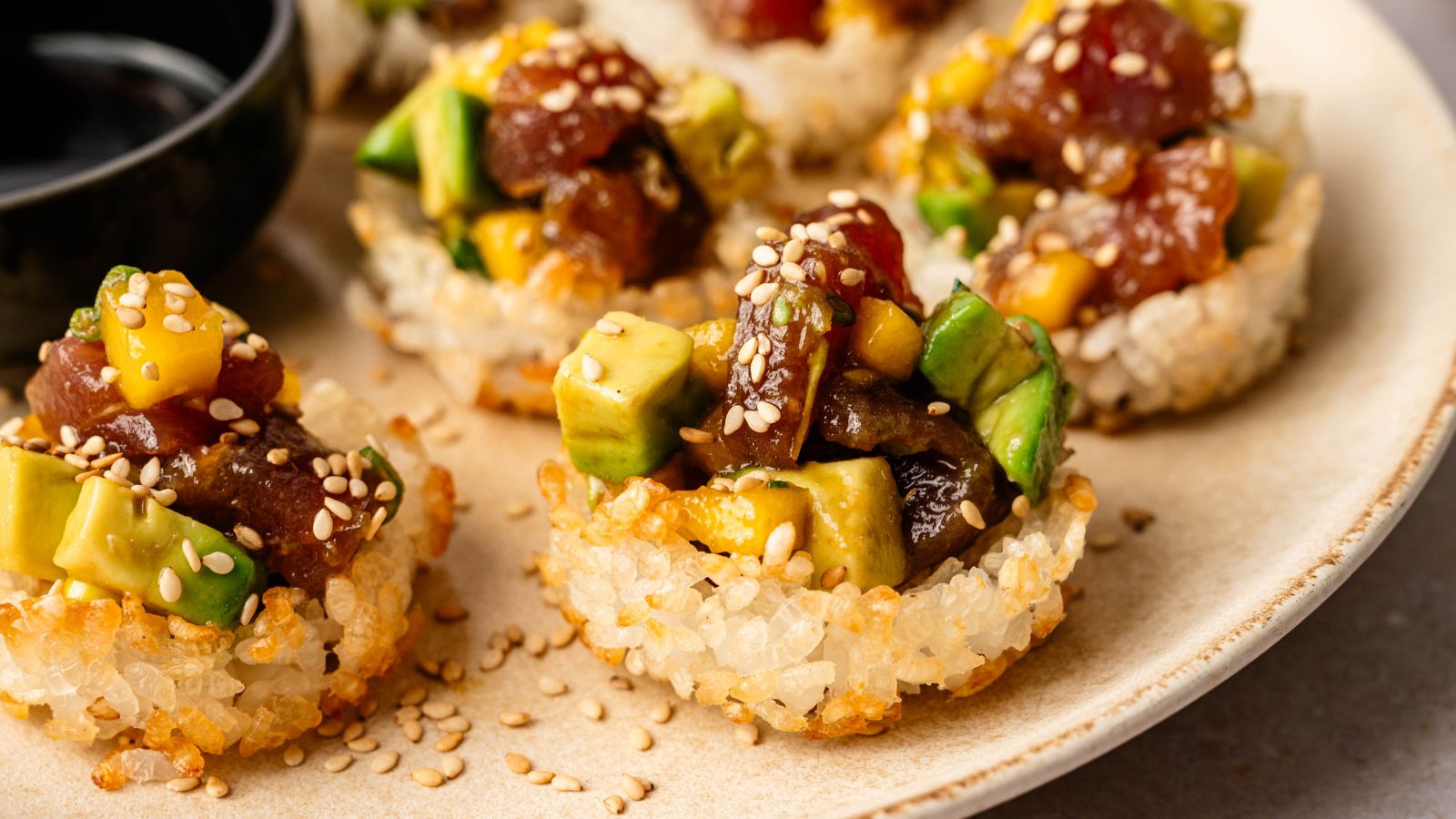 Close-up of crispy rice rounds topped with diced tuna, avocado, mango, and sesame seeds, served on a beige plate with a small dish of soy sauce.