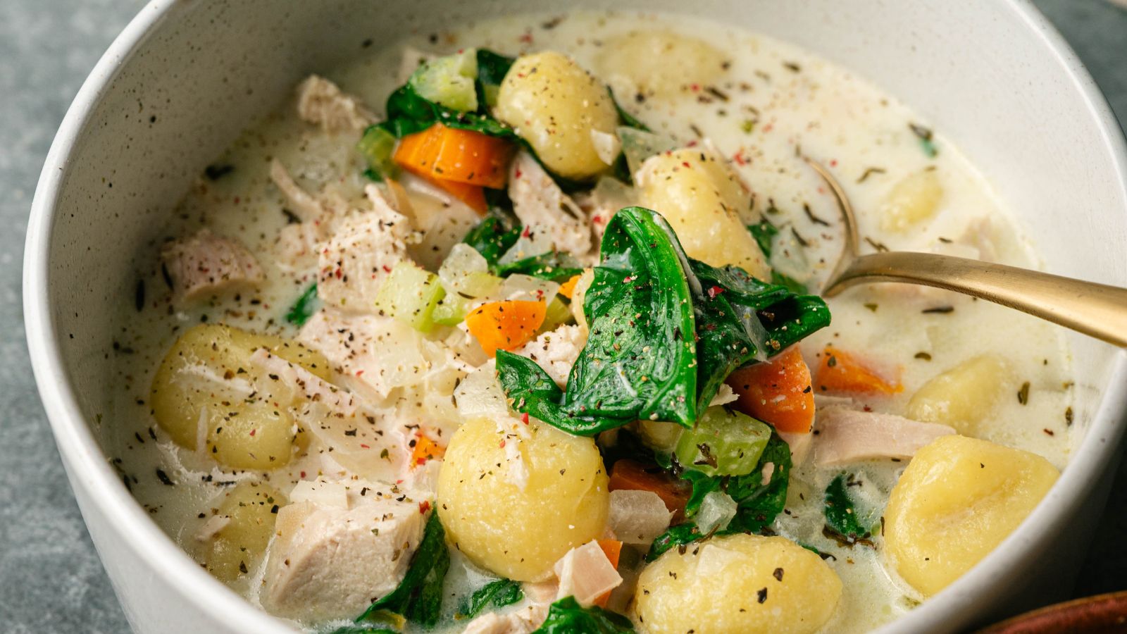 A bowl of creamy soup with gnocchi, spinach, carrots, celery, chunks of chicken, and black pepper, with a spoon inside.