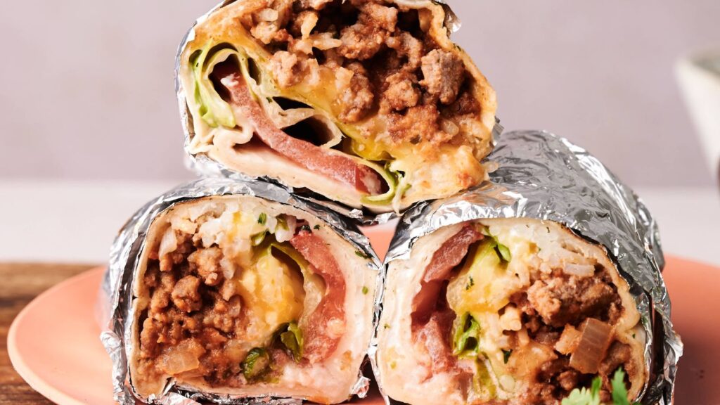 Three beef and vegetable burritos wrapped in foil are stacked on a pink plate, showing their fillings of ground beef, lettuce, tomato, and melted cheese.