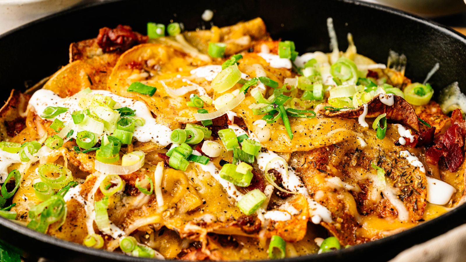 A close-up of a skillet filled with cheesy nachos topped with sliced green onions, melted cheese, sour cream, and various seasonings.
