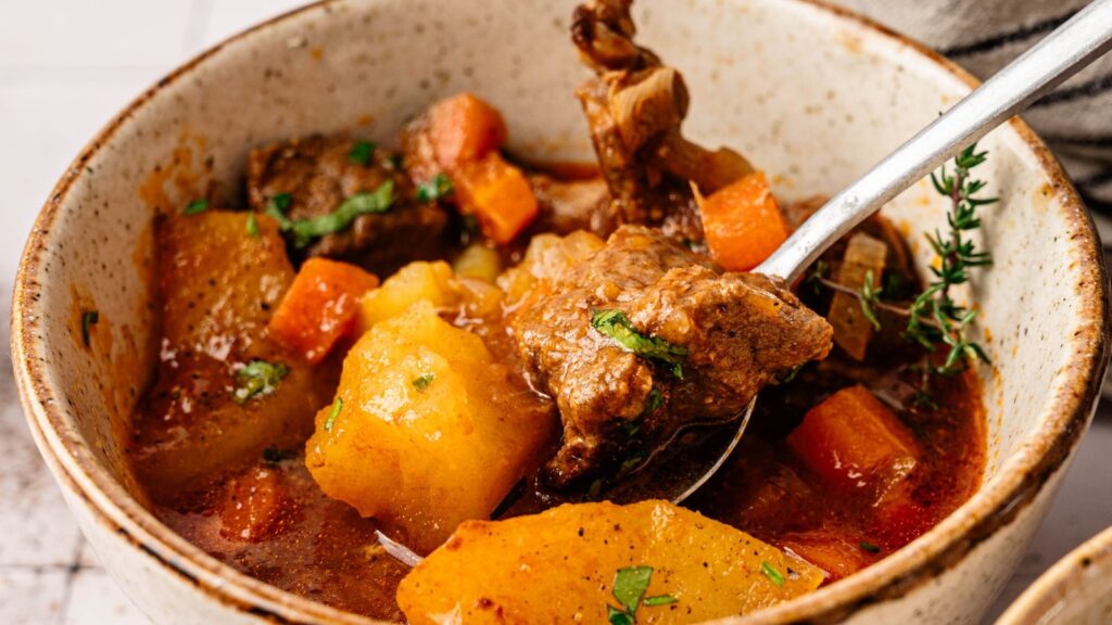 A bowl of beef stew with chunks of beef, potatoes, and carrots in a rich broth, garnished with herbs.