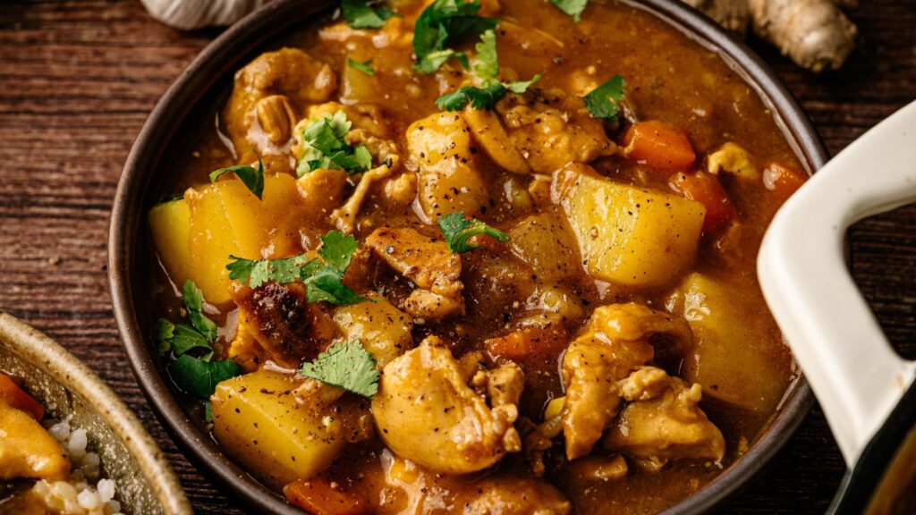 A bowl of chicken curry with potatoes, carrots, and fresh herbs, served on a wooden table.