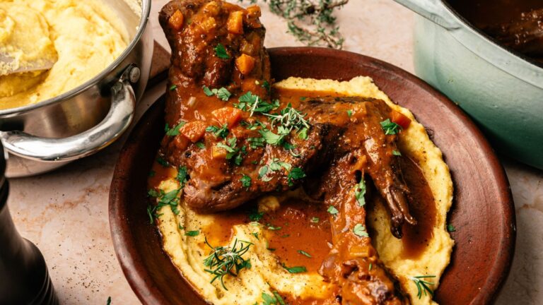 Braised lamb shanks with vegetable sauce served over creamy polenta, garnished with fresh herbs, on a brown plate next to a pot of polenta.