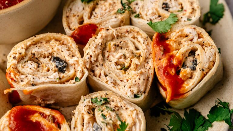 Close-up of sliced tortilla roll-ups filled with shredded chicken, herbs, and olives, some topped with red sauce, garnished with fresh cilantro.