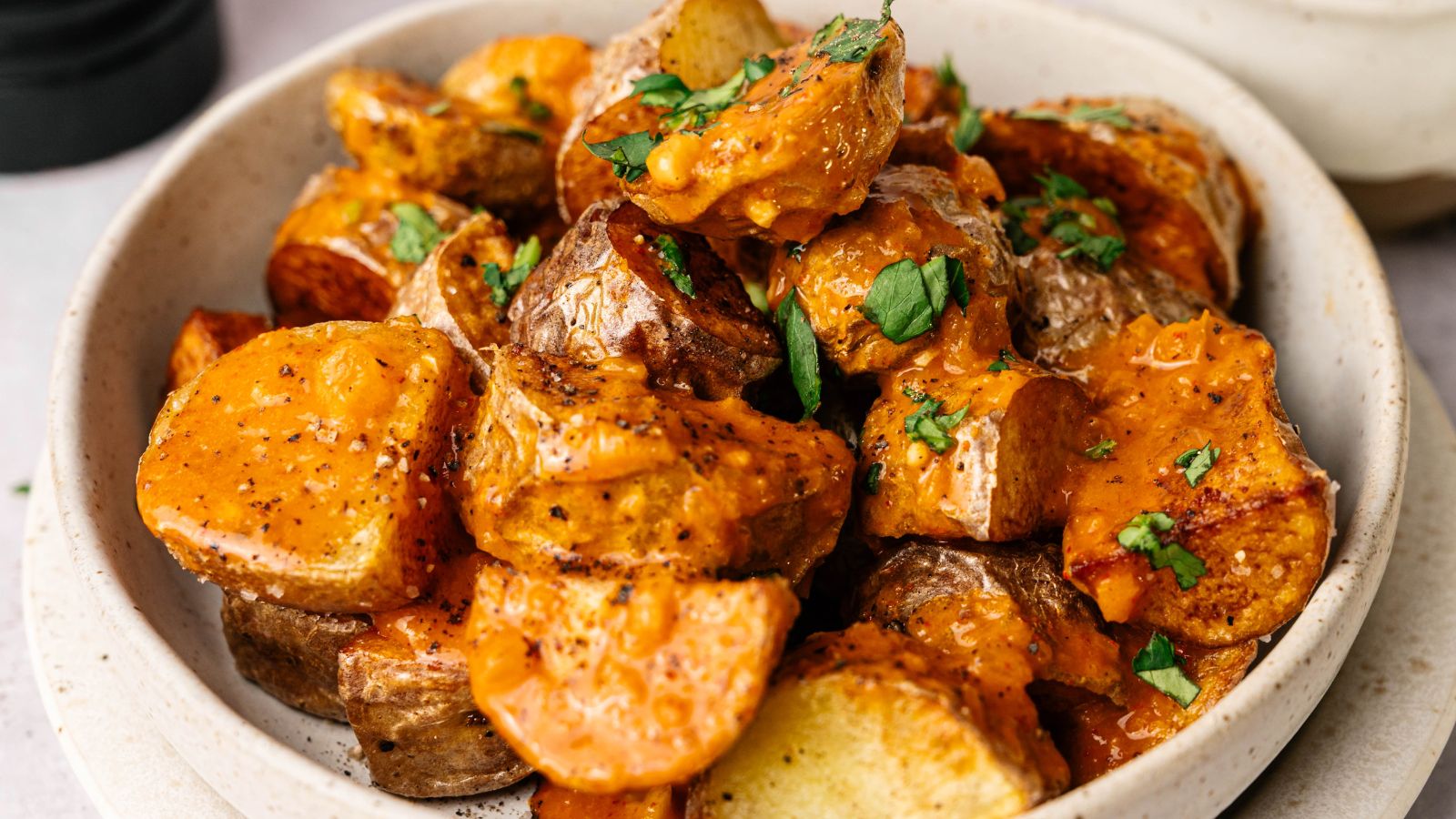 A bowl of roasted potato wedges covered in a creamy orange sauce and garnished with chopped herbs.