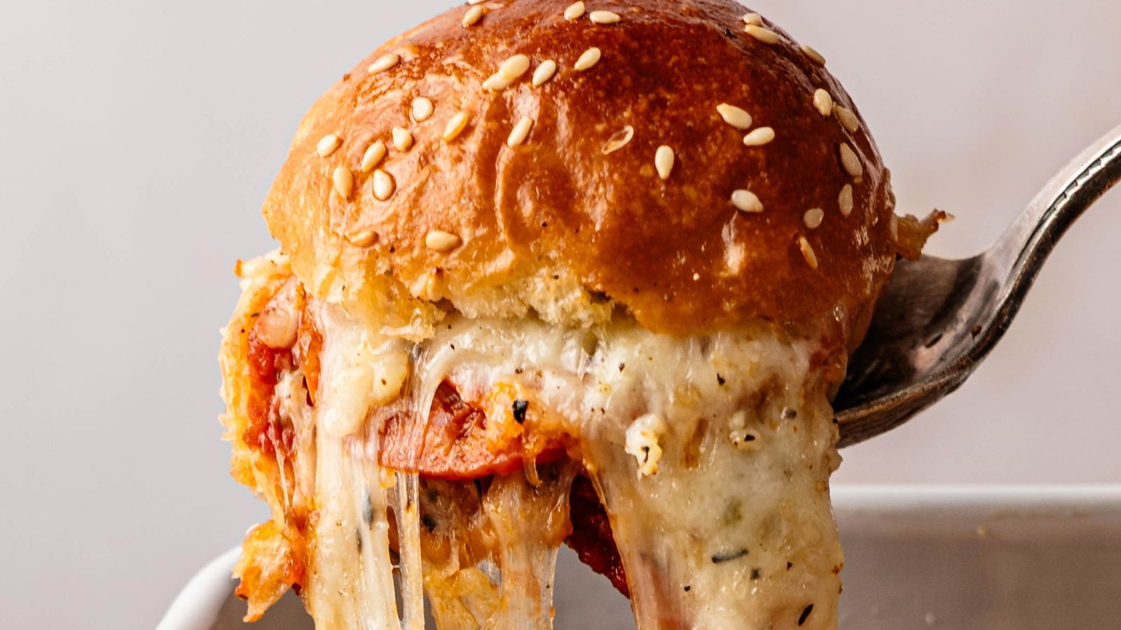 A close-up of a fork lifting a cheesy, sesame-topped slider filled with tomato sauce and melted cheese.