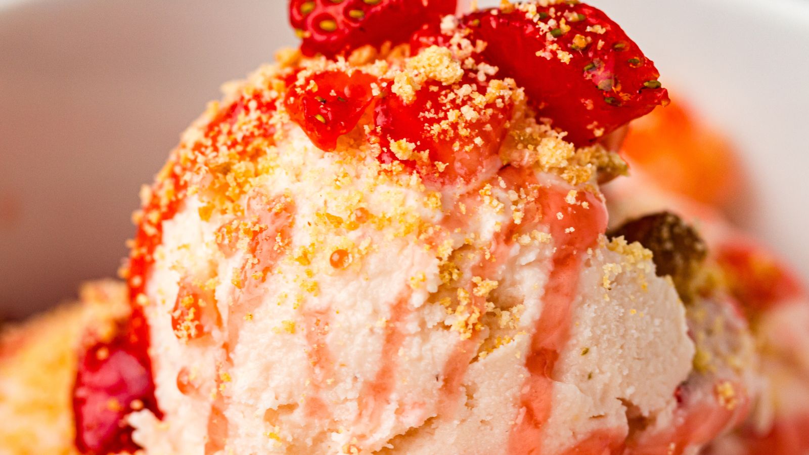 Close-up of ice cream topped with strawberry pieces, strawberry sauce, and crumbled cookie or biscuit.
