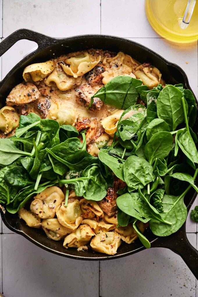 A cast iron skillet filled with Marry Me Chicken Tortellini, tender pieces of chicken, and a generous handful of fresh spinach leaves on top.