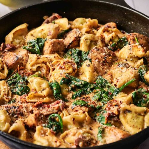 A skillet filled with Marry Me Chicken Tortellini, spinach, and chicken in a creamy sauce, with a bowl of grated cheese in the background.