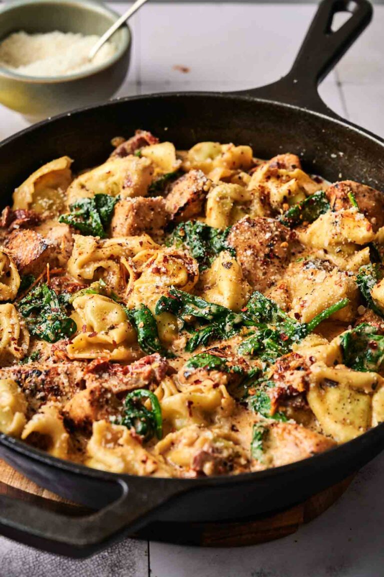 A skillet filled with Marry Me Chicken Tortellini, spinach, and chicken in a creamy sauce, with a bowl of grated cheese in the background.