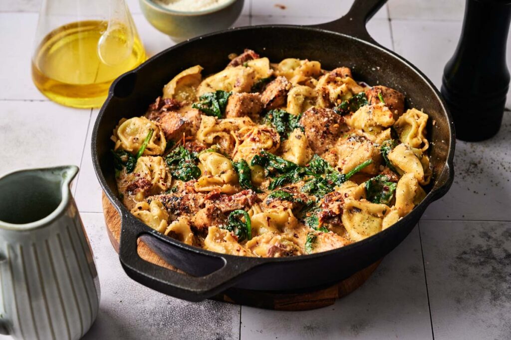 A cast iron skillet filled with Marry Me Chicken Tortellini, tender spinach, and seasoned chicken pieces, set on a white tiled surface beside a jug, oil bottle, and pepper grinder.