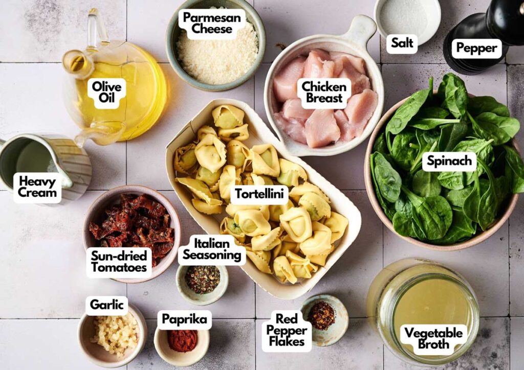 A top-down view of ingredients for Marry Me Chicken Tortellini, featuring tortellini, chicken, spinach, sun-dried tomatoes, heavy cream, seasonings, olive oil, parmesan, broth, salt, and pepper.