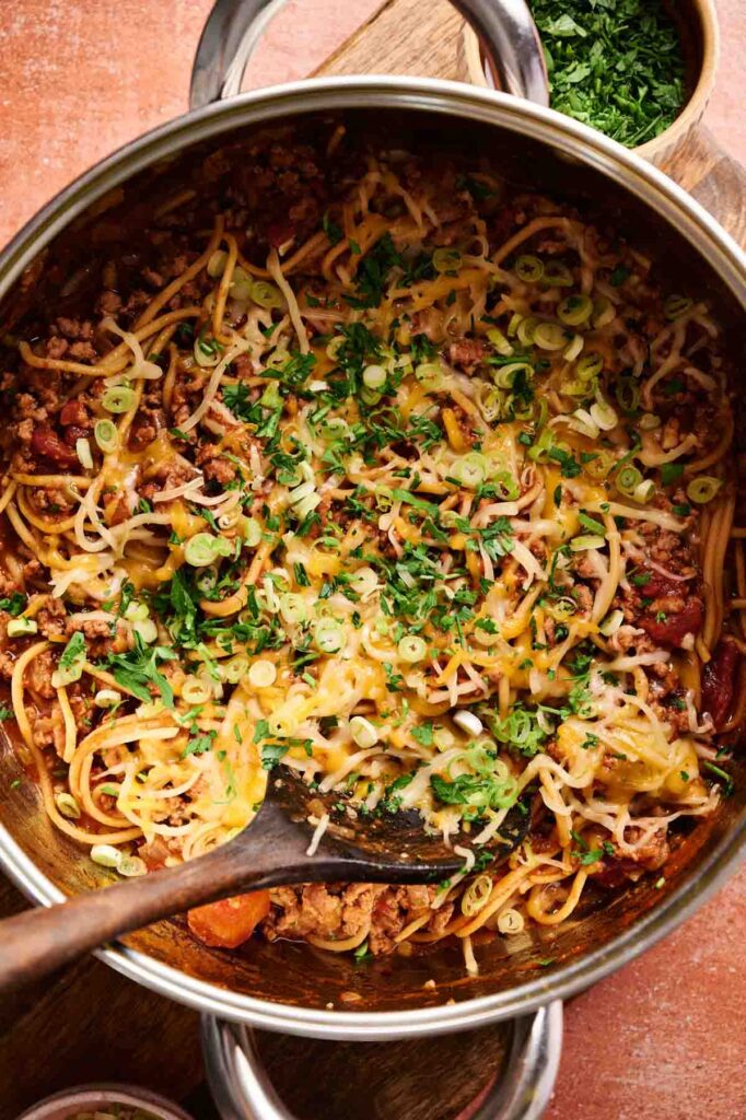 A pot of Mexican Spaghetti mixed with ground meat, beans, melted cheese, and chopped green onions, with a wooden spoon and a bowl of chopped herbs nearby.