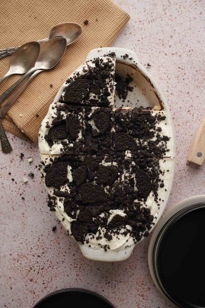 A delicious Oreo Ice Cream Cake dessert with crumbled chocolate cookies on top, partially sliced, sits next to three spoons and a cloth on a pink countertop.