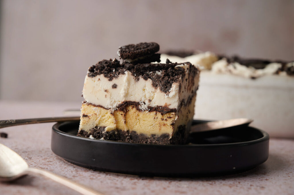 A slice of layered Oreo Ice Cream Cake with cookie crumbs and a mini cookie on top, served on a black plate with a fork and spoon.
