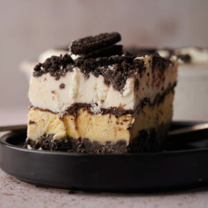 A close-up of an Oreo Ice Cream Cake dessert with cookie crumbs and cream, topped with a chocolate sandwich cookie, served on a black plate.