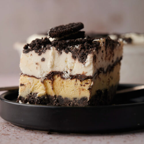 A close-up of an Oreo Ice Cream Cake dessert with cookie crumbs and cream, topped with a chocolate sandwich cookie, served on a black plate.