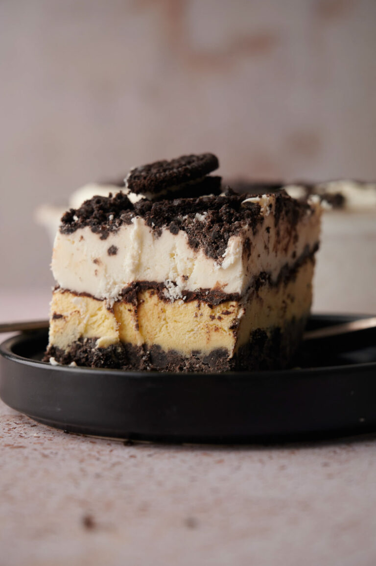 A close-up of an Oreo Ice Cream Cake dessert with cookie crumbs and cream, topped with a chocolate sandwich cookie, served on a black plate.