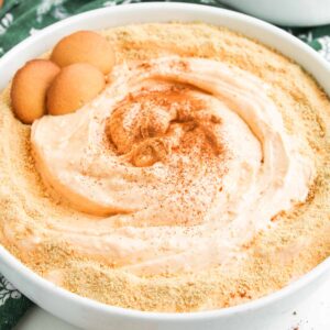 A dish of Pumpkin Pie Dip topped with graham cracker crumbs and cinnamon, garnished with vanilla wafer cookies, sits on a green patterned cloth.