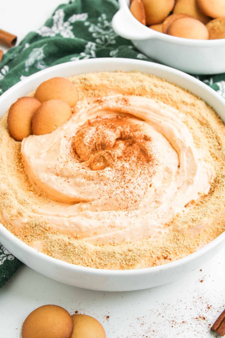 A dish of Pumpkin Pie Dip topped with graham cracker crumbs and cinnamon, garnished with vanilla wafer cookies, sits on a green patterned cloth.