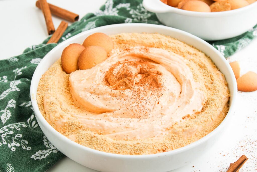 A bowl of creamy Pumpkin Pie Dip topped with graham cracker crumbs and cinnamon, garnished with vanilla wafer cookies, sits on a green patterned cloth.