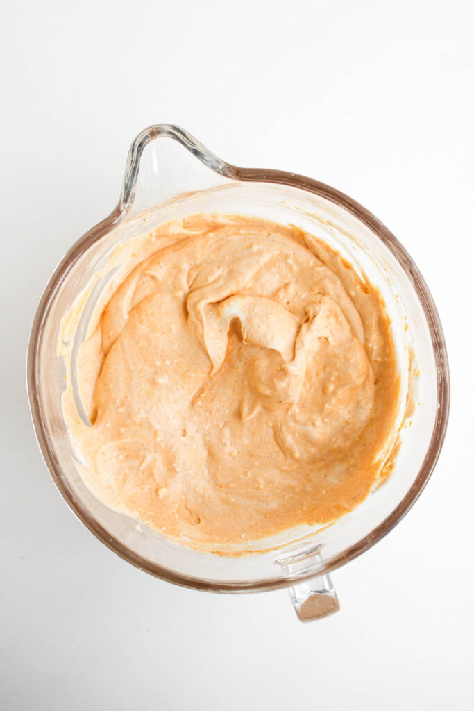 A glass mixing bowl containing a thick, creamy orange Pumpkin Pie Dip batter on a white surface.