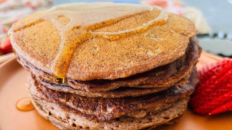 A stack of three pancakes with syrup drizzled on top, served on a plate with sliced strawberries on the side.