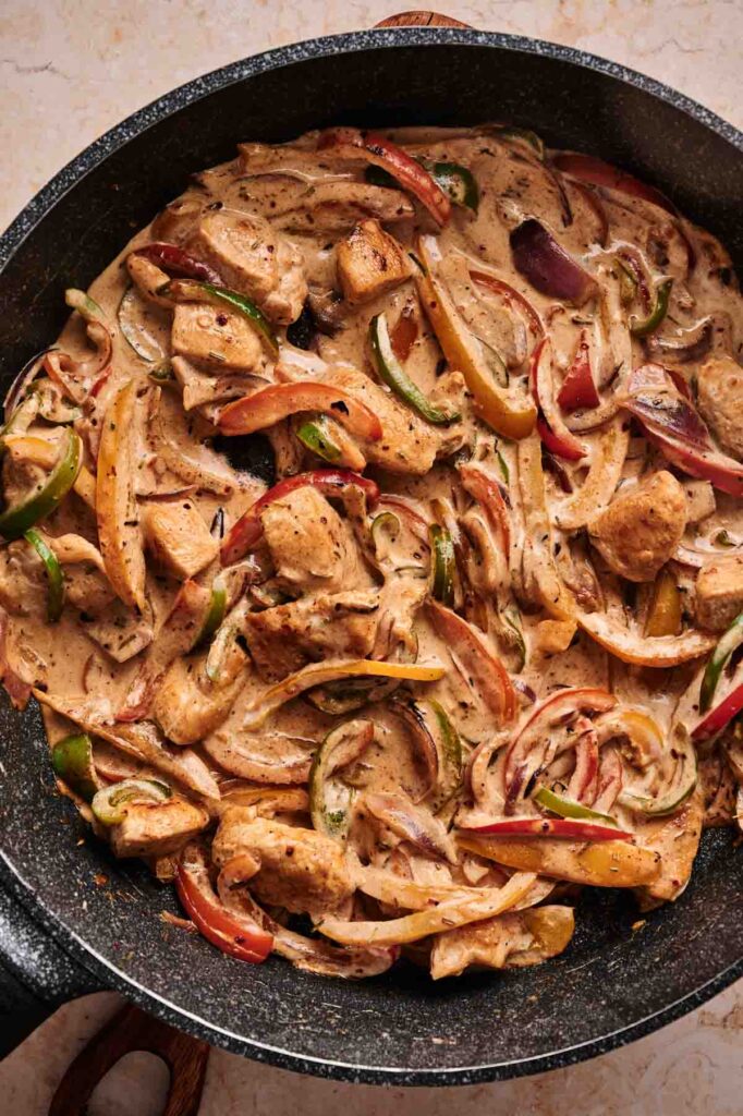 A skillet filled with creamy cooked chicken pieces, sliced red and green bell peppers, onions, seasonings, and a touch of Rasta Pasta flair.