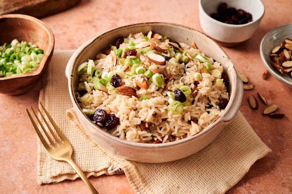 A bowl of Rice Pilaf topped with sliced almonds, dried cranberries, and chopped green onions, with a gold fork beside it on a beige napkin.
