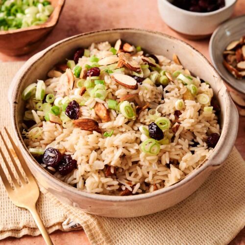 A bowl of Rice Pilaf topped with sliced almonds, dried cranberries, and chopped green onions sits on a beige napkin, with a fork and small bowls of ingredients nearby.