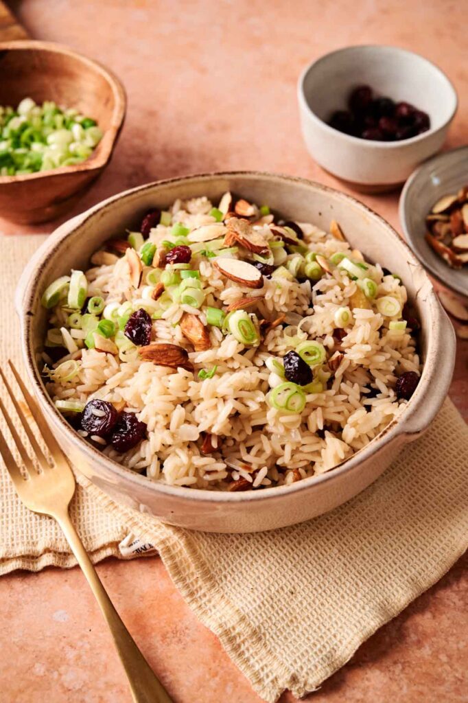 A bowl of Rice Pilaf topped with sliced almonds, dried cranberries, and chopped green onions sits on a beige napkin, with a fork and small bowls of ingredients nearby.