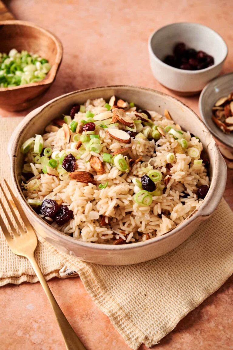 A bowl of Rice Pilaf topped with sliced almonds, dried cranberries, and chopped green onions sits on a beige napkin, with a fork and small bowls of ingredients nearby.