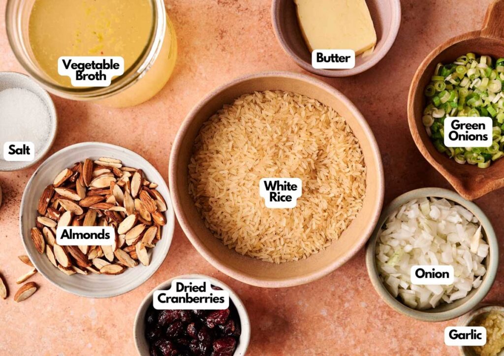 A variety of labeled ingredients for Rice Pilaf are arranged on a table, including white rice, vegetable broth, butter, green onions, onion, garlic, salt, almonds, and dried cranberries.
