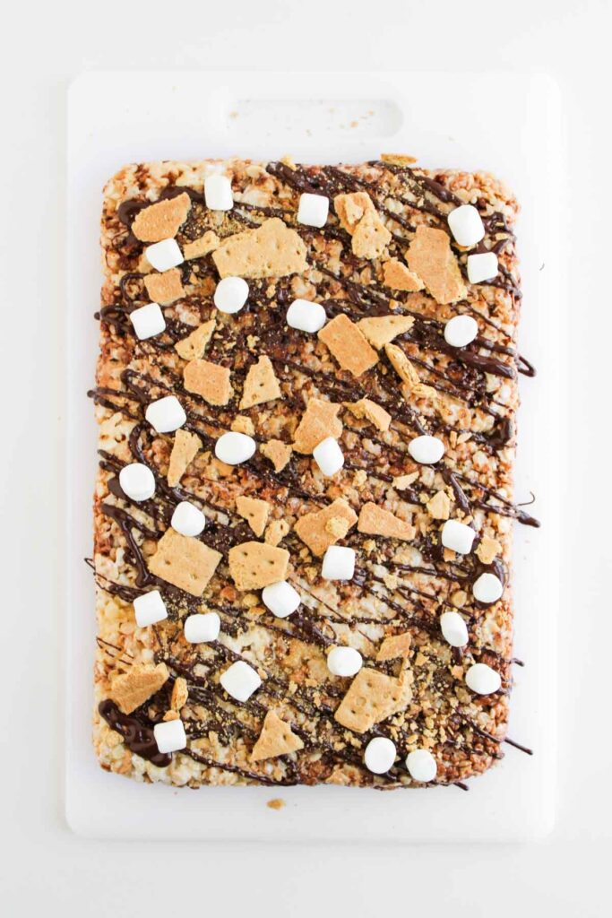 A rectangular S'mores Rice Krispies treat topped with mini marshmallows, chocolate drizzle, and pieces of graham crackers sits on a white cutting board.