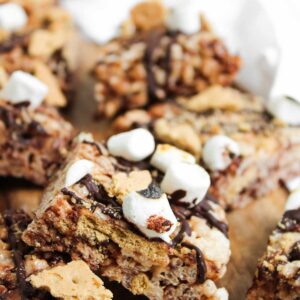 Close-up of S'mores Rice Krispies dessert bars made with crispy rice cereal, mini marshmallows, chocolate drizzle, and pieces of graham crackers on a wooden surface.