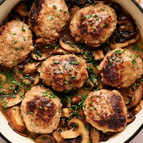 Six Salisbury Steak patties, browned to perfection, rest atop sliced mushrooms in a creamy sauce, garnished with chopped chives in a white skillet.