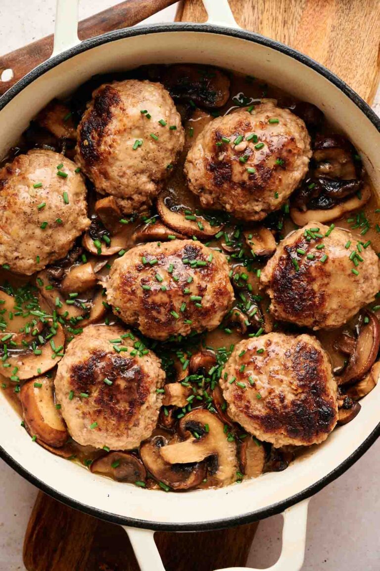 Six Salisbury Steak patties, browned to perfection, rest atop sliced mushrooms in a creamy sauce, garnished with chopped chives in a white skillet.