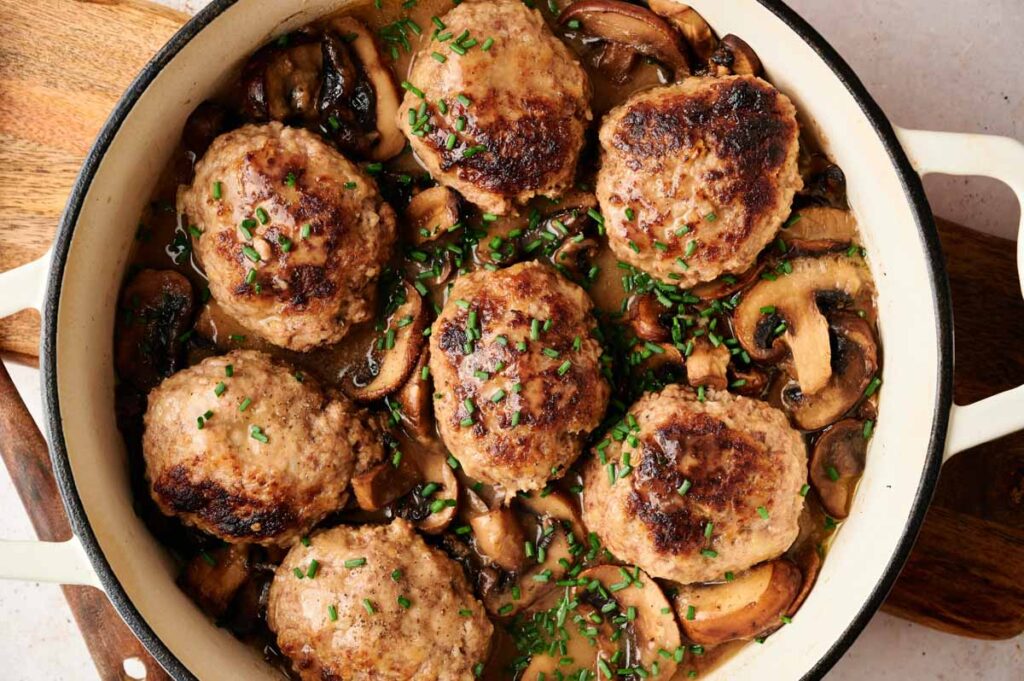 Large Salisbury Steak meatballs in a creamy mushroom sauce are arranged in a round white pan, garnished with chopped chives.
