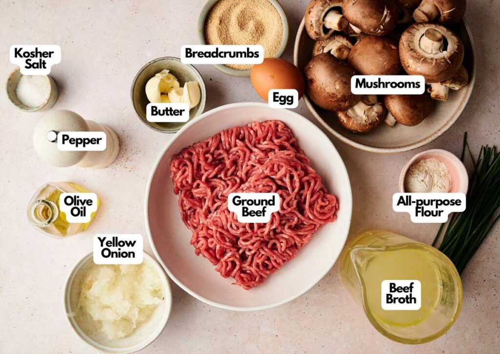 A top view of labeled Salisbury Steak ingredients for a recipe, including ground beef, mushrooms, egg, breadcrumbs, all-purpose flour, beef broth, yellow onion, olive oil, butter, pepper, and kosher salt.