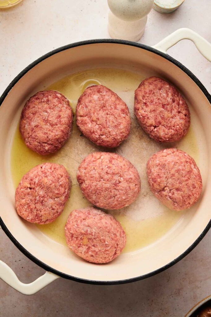 Seven raw beef patties, perfect for Salisbury Steak, are arranged in a white pan with oil, ready to be cooked.