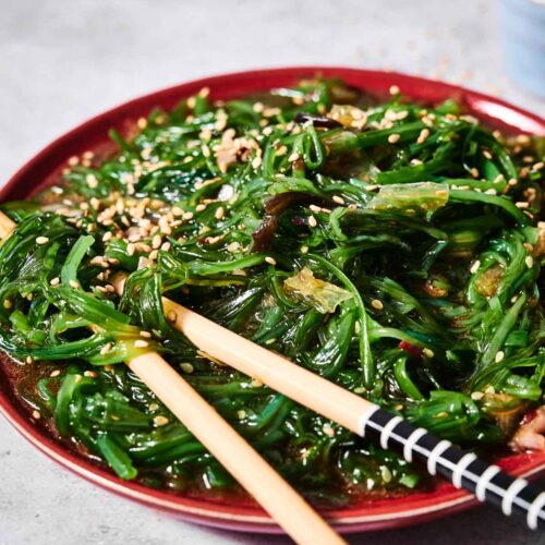 A fresh plate of Seaweed Salad topped with sesame seeds, served with a pair of chopsticks on a gray surface.