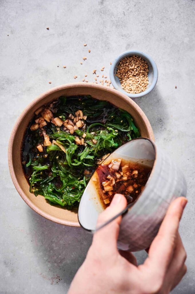 A hand pours dressing over a bowl of Seaweed Salad, with a small dish of sesame seeds beside it on a light gray surface.