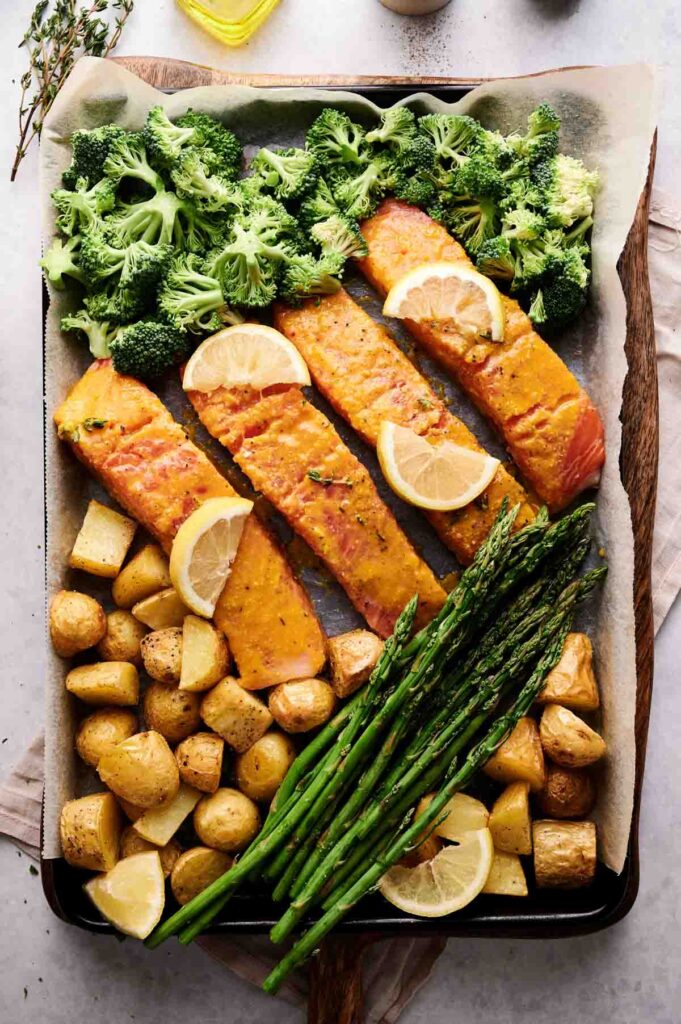 Sheet Pan Salmon features seasoned fillets topped with lemon slices, surrounded by broccoli, asparagus, and roasted potato cubes on a baking sheet.